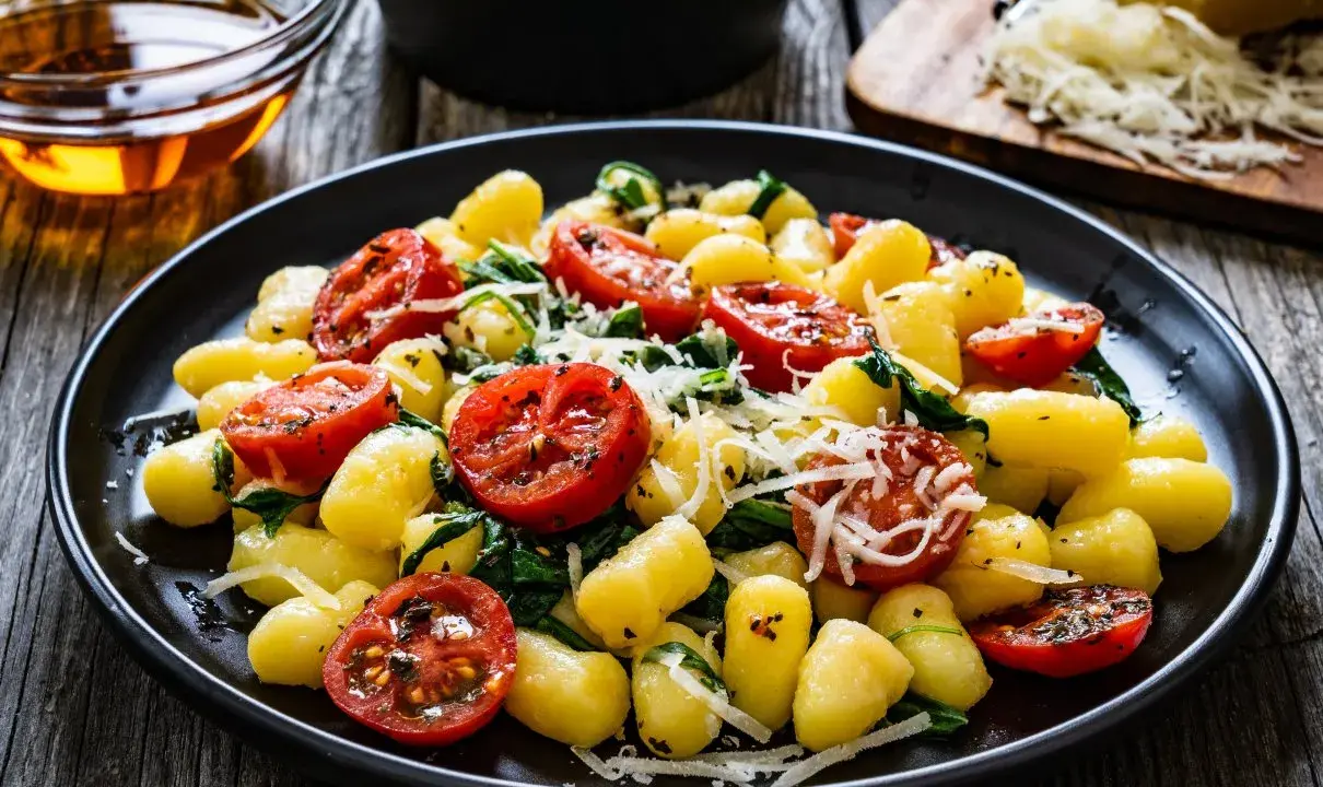 Gnocchi ze szpinakiem i pomidorkami koktajlowymi, posypane parmezanem, na czarnym talerzu.