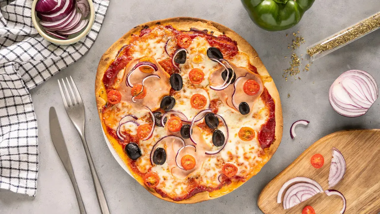 Gotowe ciasto na pizzę z szynką, oliwkami, pomidorkami i czerwoną cebulą. Obok sztućce i miseczka z cebulą.