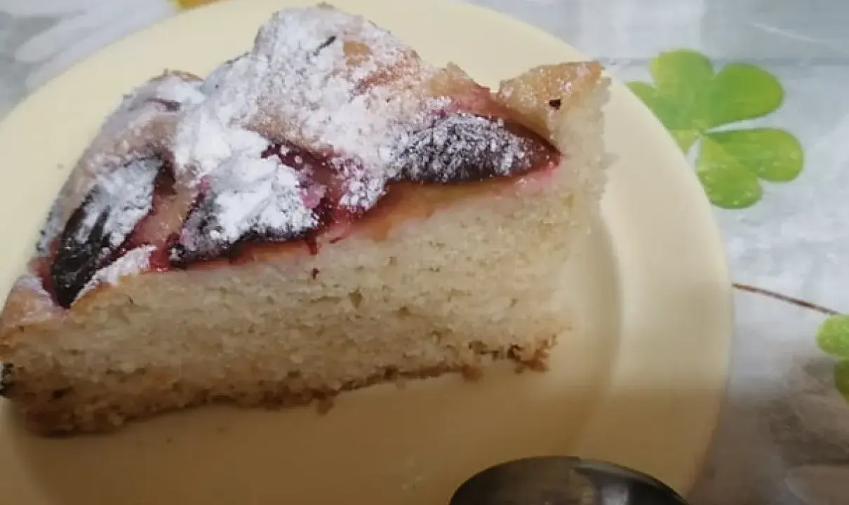 Pyszne ciasto z mrożonymi śliwkami, posypane cukrem pudrem, na żółtym talerzyku. Idealne na deser.