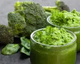 Dwa słoiczki z zielonym puree z brokułów, ozdobione natką pietruszki, na tle świeżych brokułów i liści szpinaku.