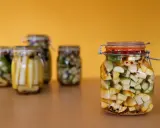 Słoiki z marynowaną cukinią, czosnkiem i przyprawami na żółtym tle.
