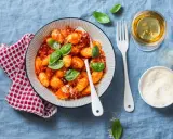 Pyszne kluski gnocchi w sosie pomidorowym, udekorowane bazylią. Obok kieliszek białego wina i miseczka parmezanu.