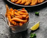 Frytki z batatów w metalowym koszyku, ozdobione gałązką tymianku, obok ćwiartki limonki i natki pietruszki.