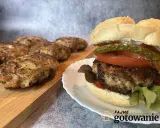 Pyszny burger wieprzowy z dodatkami na talerzyku, obok leżą świeżo usmażone kotlety.