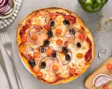 Gotowe ciasto na pizzę z szynką, oliwkami, pomidorkami i czerwoną cebulą. Obok sztućce i miseczka z cebulą.