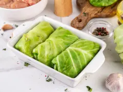 Gotowe do pieczenia gołąbki z młodej kapusty w naczyniu żaroodpornym, obok składniki: mięso mielone, czosnek, przyprawy.