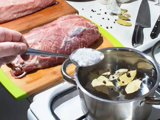 Przygotowanie solanki do wędzenia. Łyżka soli trafia do garnka z wodą, liśćmi laurowymi i pieprzem, obok surowego mięsa.