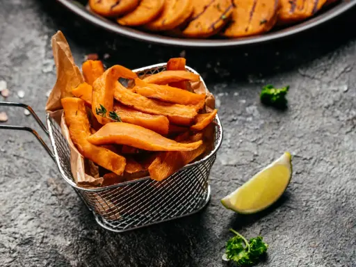 Frytki z batatów w metalowym koszyku, ozdobione gałązką tymianku, obok ćwiartki limonki i natki pietruszki.