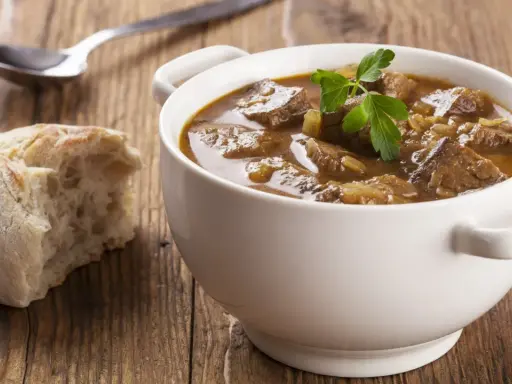 Aromaticzny, tradycyjny gulasz wołowy w białej miseczce, z gałązką pietruszki, obok kromka chleba i łyżka.