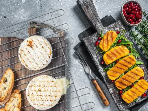 Dwa okrągłe sery camembert grillowane na ruszcie, obok pieczywo i grillowane kawałki sera na desce z granatem.