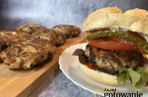 Pyszny burger wieprzowy z dodatkami na talerzyku, obok leżą świeżo usmażone kotlety.