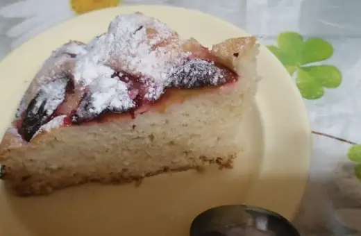 Pyszne ciasto z mrożonymi śliwkami, posypane cukrem pudrem, na żółtym talerzyku. Idealne na deser.