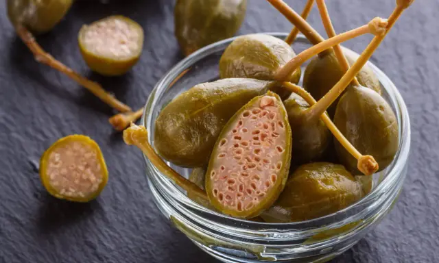 Kapary w szklanej miseczce, jeden przekrojony, na ciemnym tle.