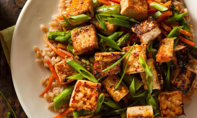 Talerz pełen brązowego ryżu z grillowanym tofu, szparagami i marchewką, posypany szczypiorkiem.