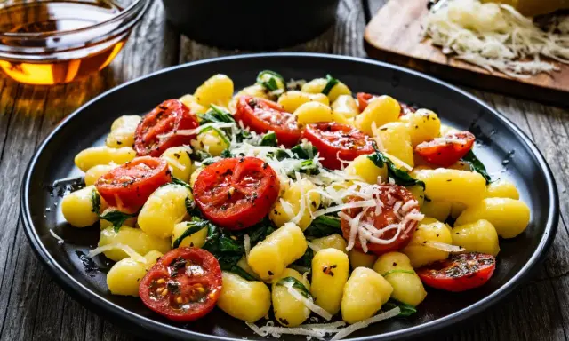 Gnocchi ze szpinakiem i pomidorkami koktajlowymi, posypane parmezanem, na czarnym talerzu.