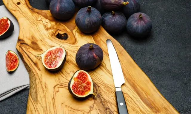 Kilka dojrzałych, ciemnofioletowych fig, niektóre przekrojone na pół, leżą na drewnianej desce obok noża.