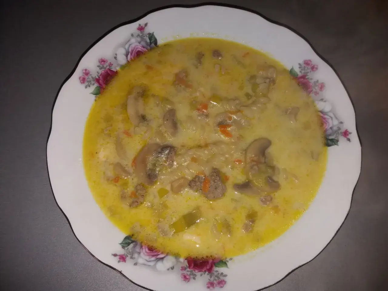 Gęsta zupa porowa z serkiem topionym, mięsem mielonym, pieczarkami i makaronem, podana na talerzu z kwiatowym wzorem.