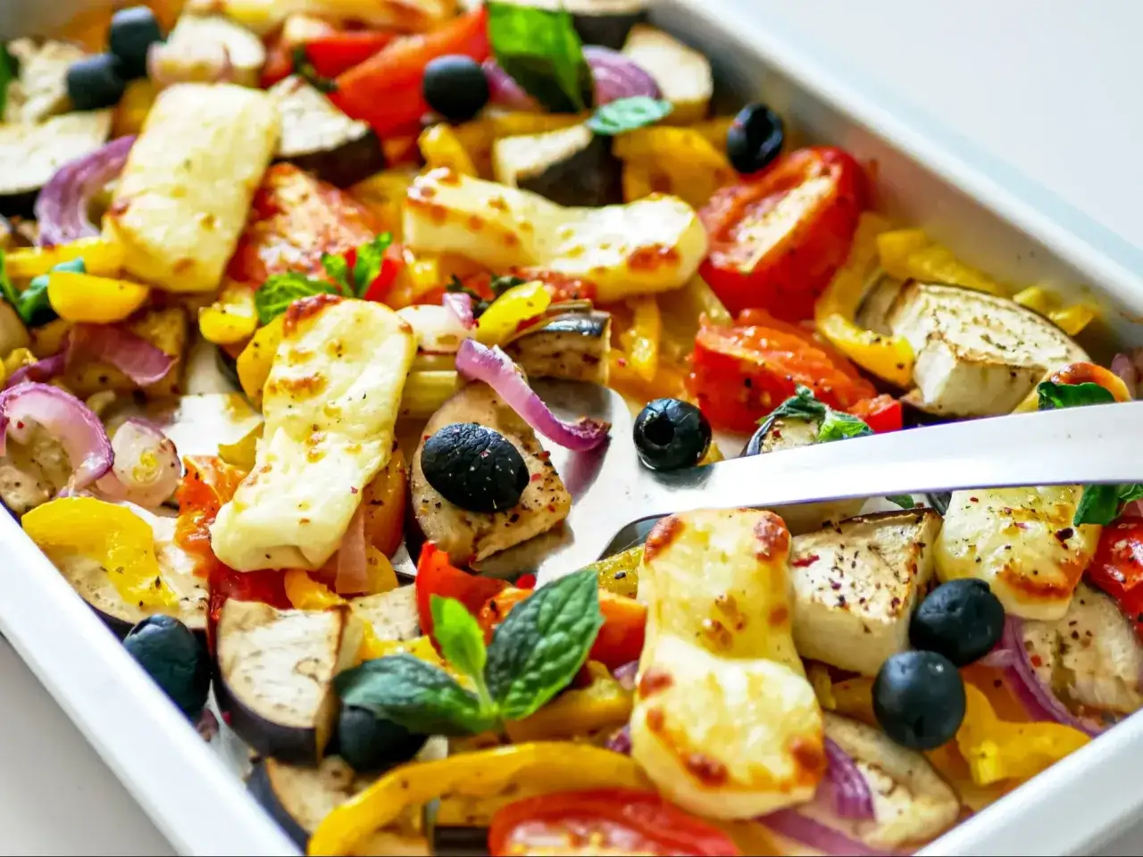 Kolorowe, pieczone warzywa z piekarnika: cukinia, papryka, pomidory, cebula i oliwki, z kawałkami sera halloumi.