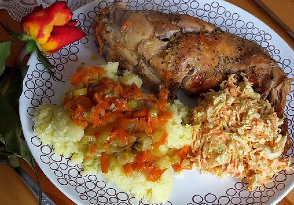 Pieczony comber z królika z puree ziemniaczanym, warzywami i surówką z marchewki. Obok leży czerwono-żółta róża.